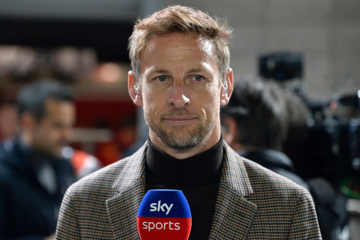 Button in high-neck dark top and beige blazer in the F1 paddock holding a Sky Sports microphone