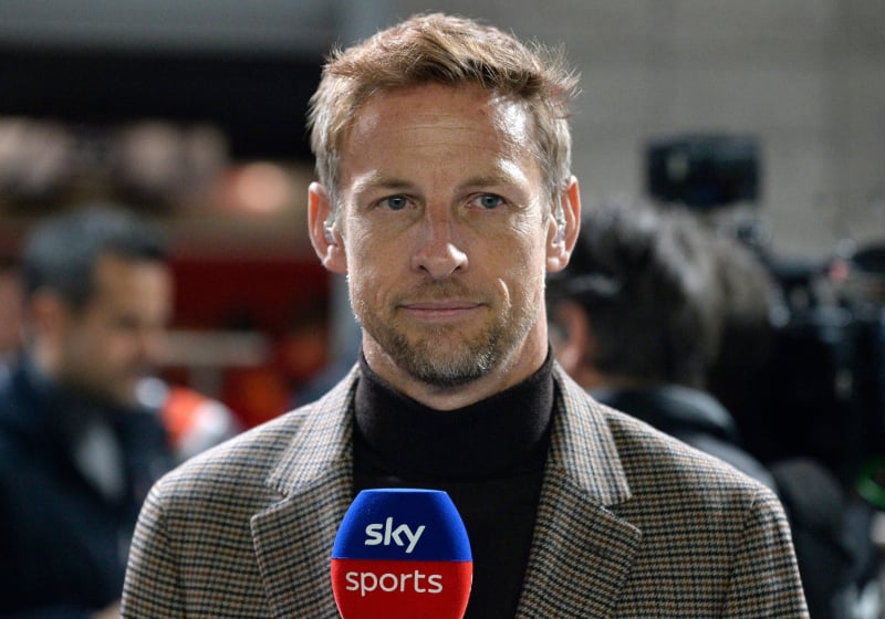 Button in high-neck dark top and beige blazer in the F1 paddock holding a Sky Sports microphone